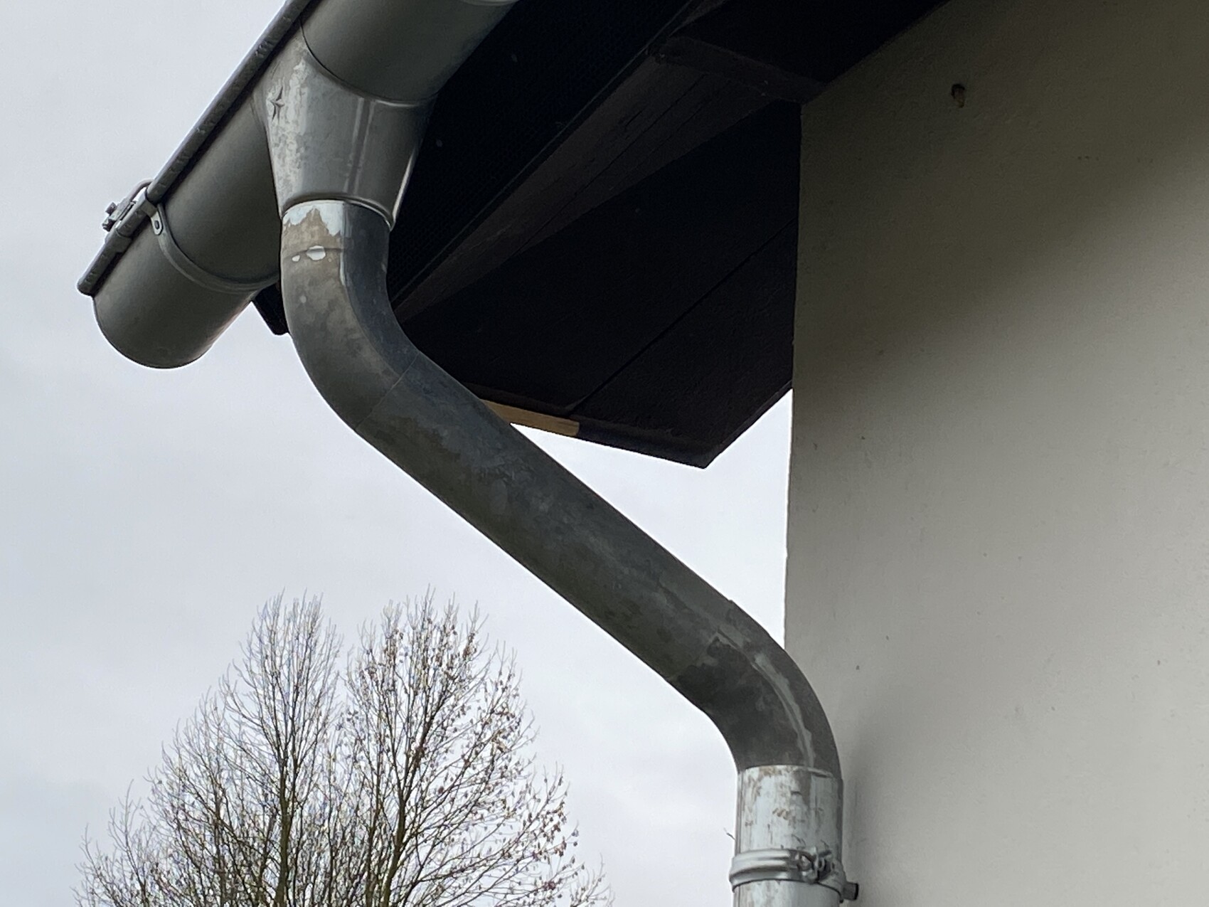 Metal gutter with bent downspout on the house, exterior view, tree in the background.