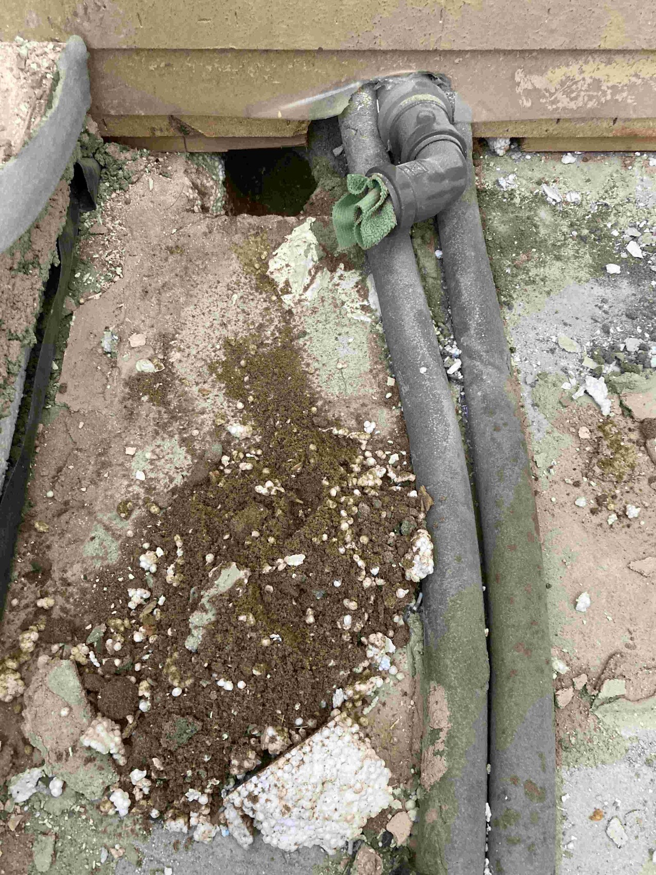 Open hole in masonry next to two pipes; soil, debris, moss, and a green cloth on the pipe.
