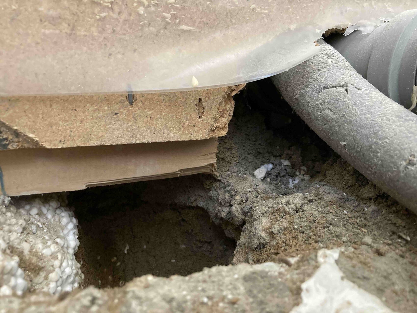 Open earth hole under a floor slab, with sand, wood scraps, and a pipe beside it.