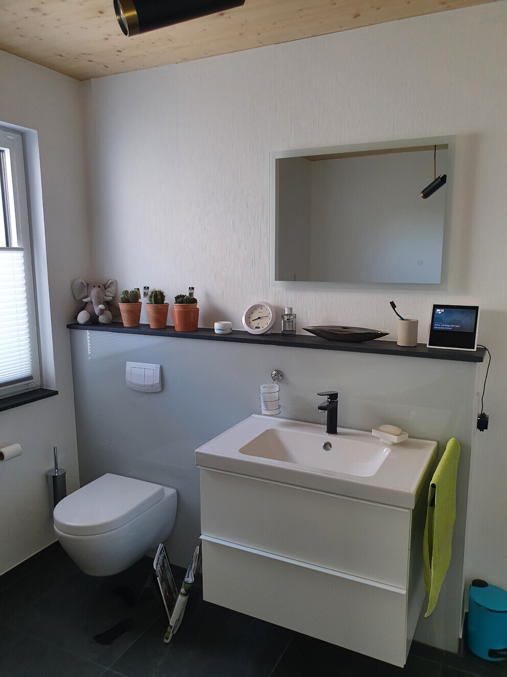 Modern bathroom with wall-mounted toilet, white washbasin, mirror, plant corner, and green towel
