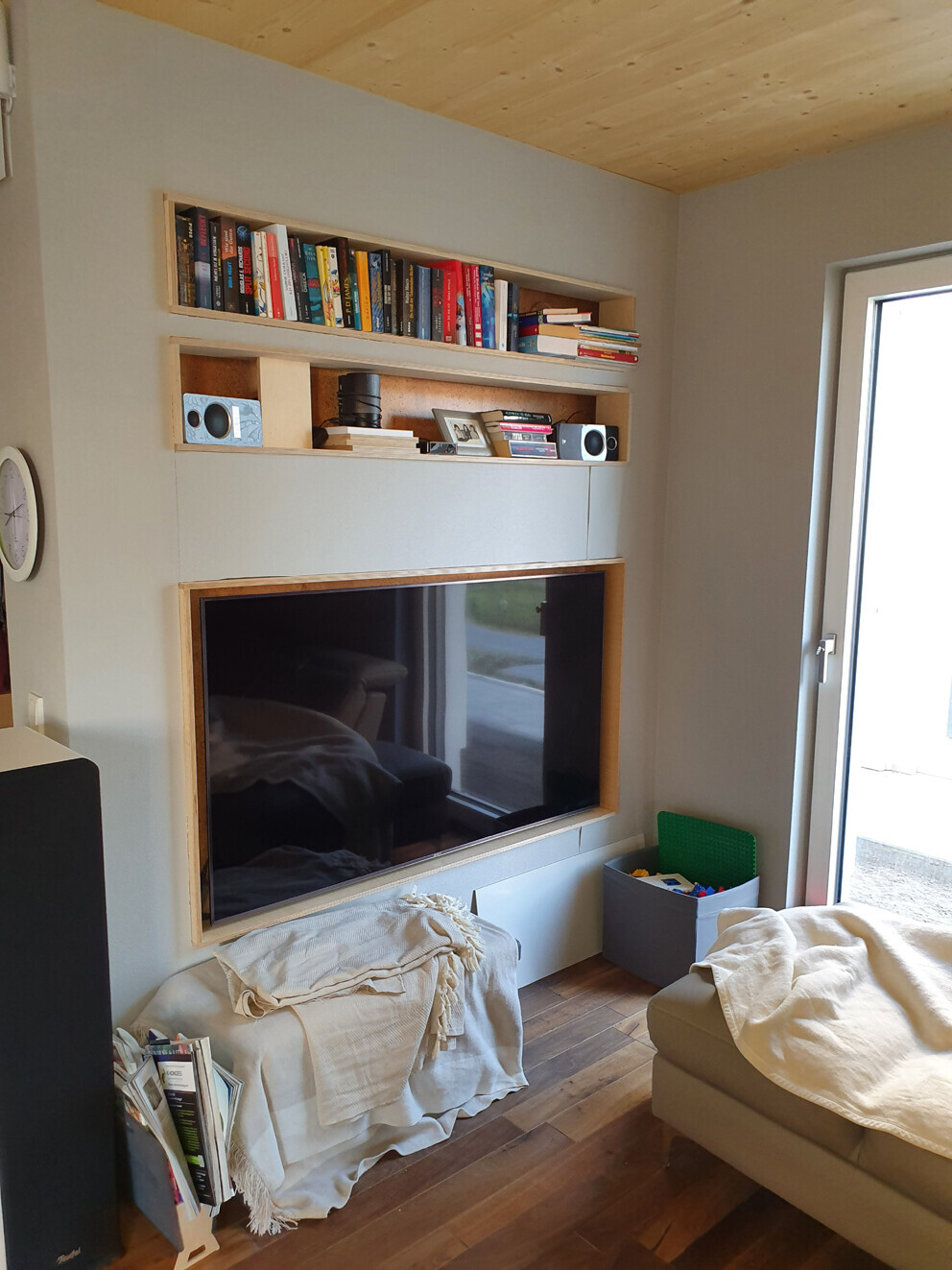 Living room with wall-mounted TV in wooden frame, bookshelves above; sofa with throw blanket.