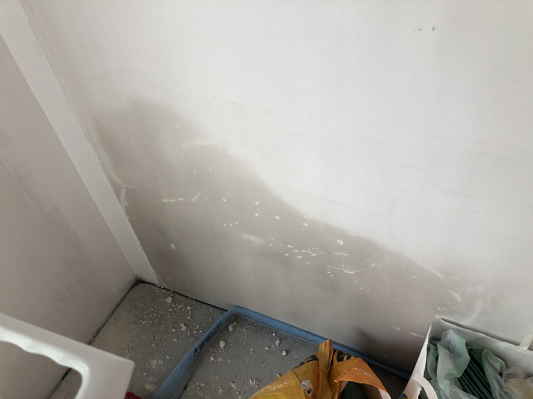Ecke eines Baustellenbereichs: weiße Wand mit Putzflecken, staubiger Boden, Bauabfälle in der Ecke.