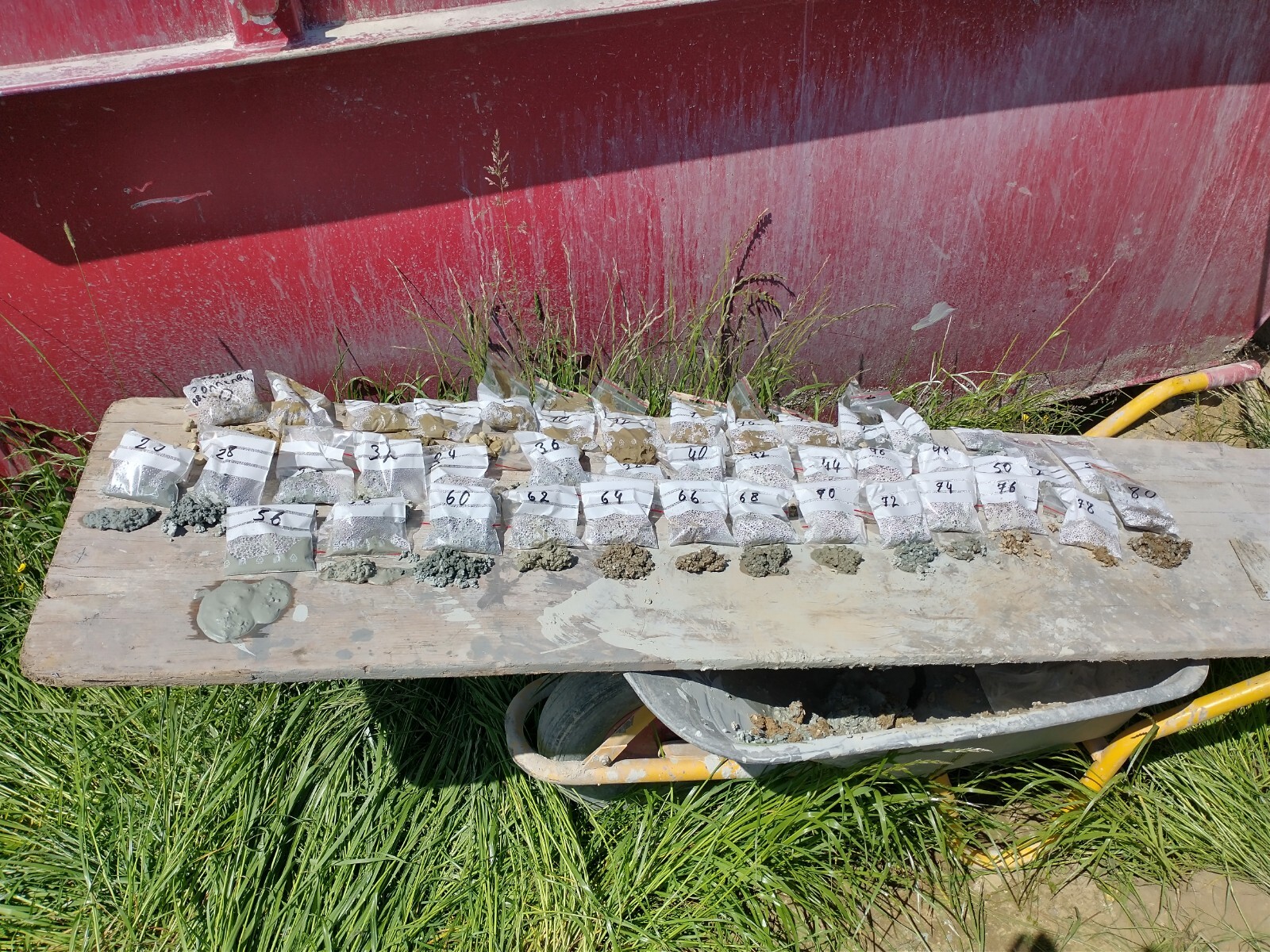 Numerous silver sample bags with numbers on a wooden board in front of a red wall.