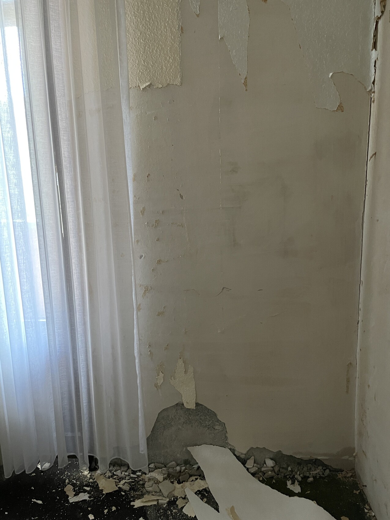 Corner of a wall with peeling paint, plaster broken off at the bottom, rubble on the floor.