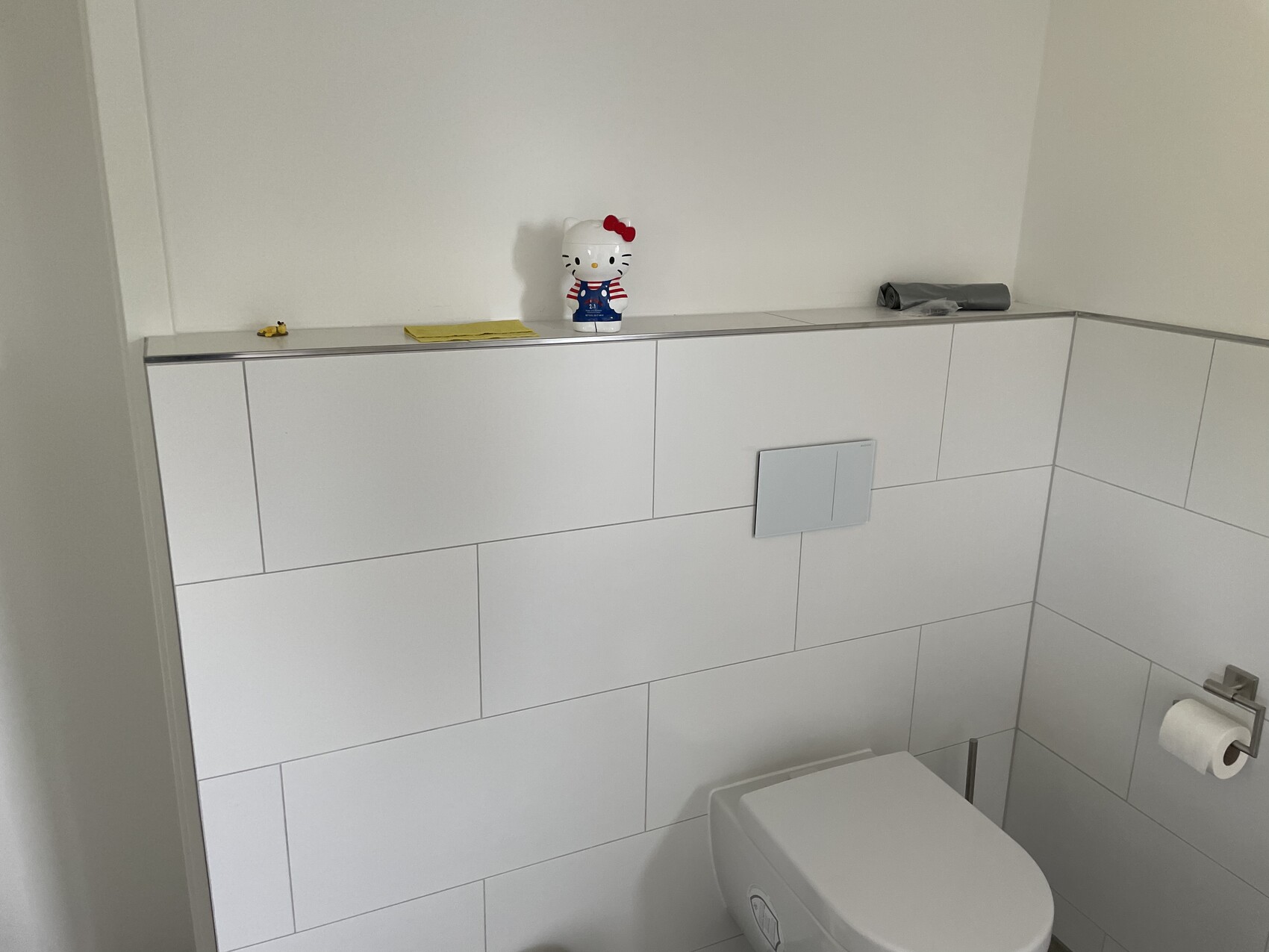 Hello Kitty-Figur auf Ablage über Toilette in hellem, weiß gefliestem Badezimmer.