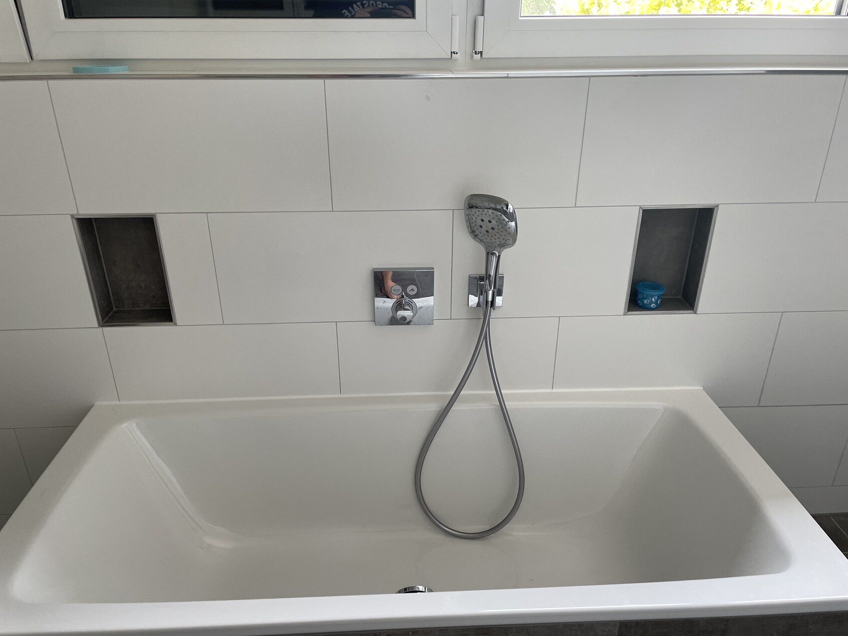 Weiße Badewanne mit Handbrause an Fliesenwand, zwei Nischen; rechte Nische enthält Becher.