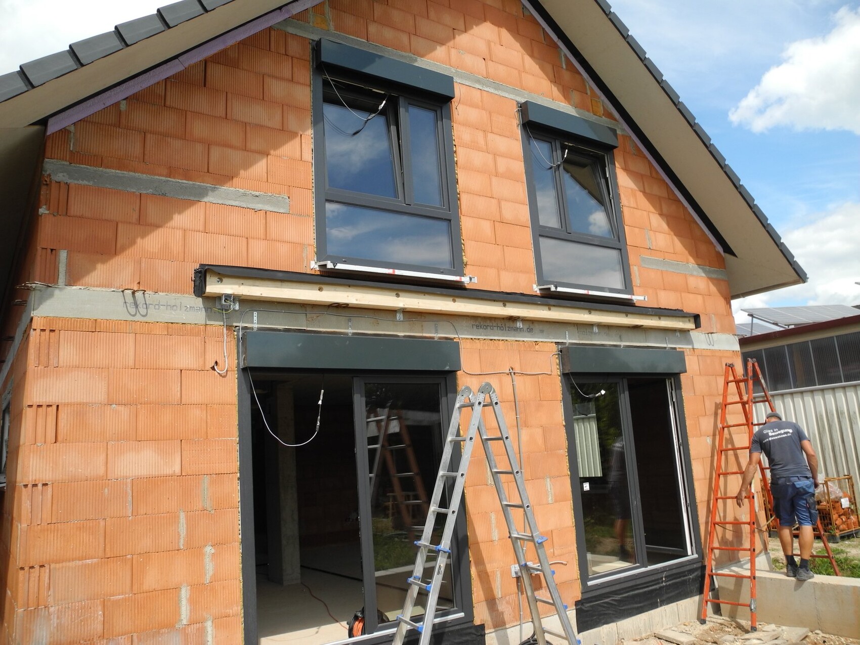 Rohbau eines zweistöckigen Hauses aus roten Ziegeln mit großen Fenstern; Leitern und Arbeiter.
