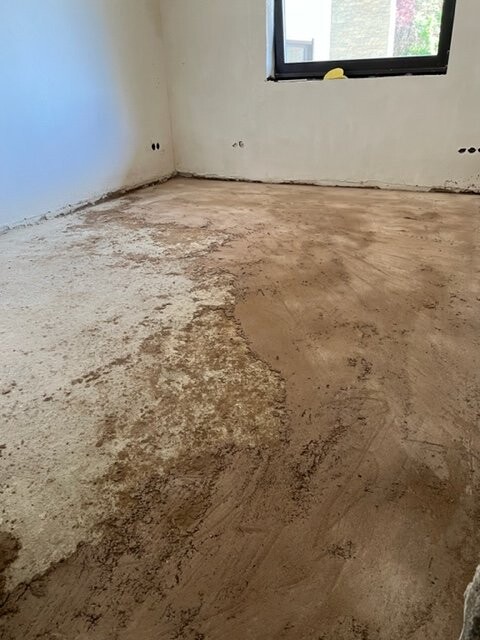 Unrenovated room with raw concrete floor, stained areas, white walls, and a window.