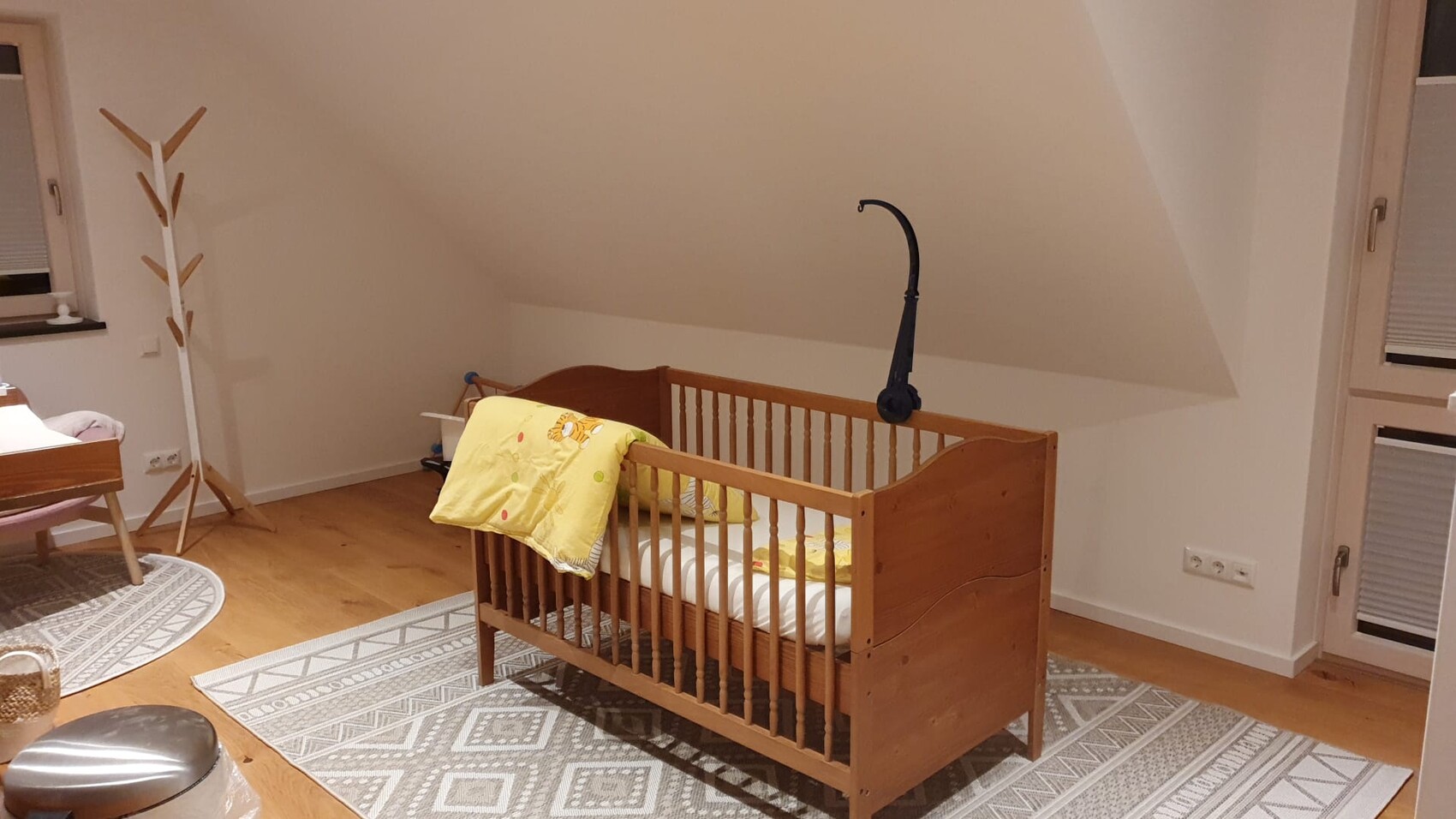 Holz-Kinderbett mit gelber Decke, graue Teppiche, Kleiderständer links, Dachschräge rechts.