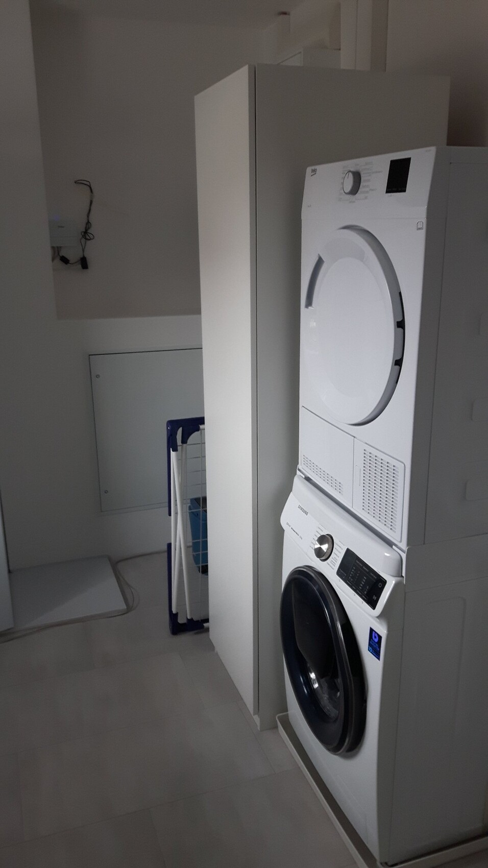 Stacked washing machine at the bottom and dryer on top in a white laundry room with cabinet.