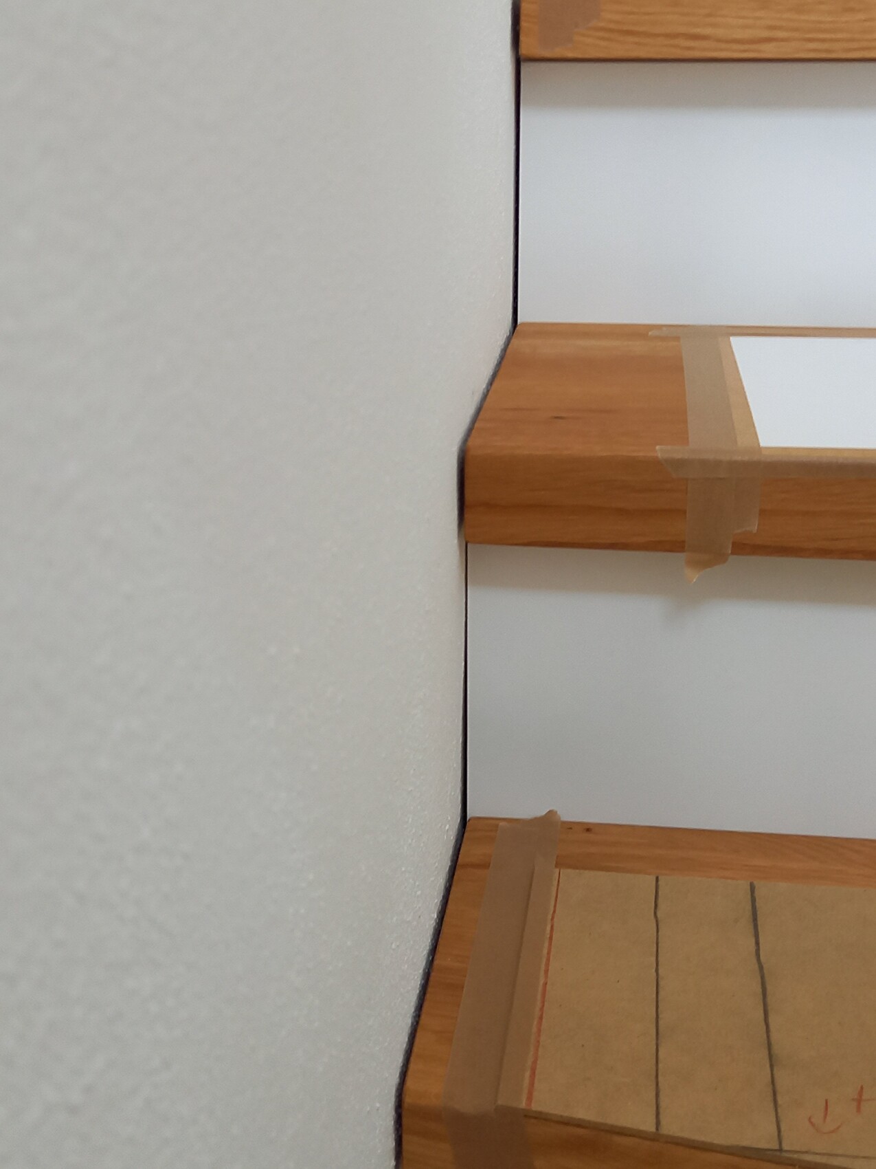 Wood staircase with tape on the edges; cardboard covering on the bottom step; gray wall on the left.