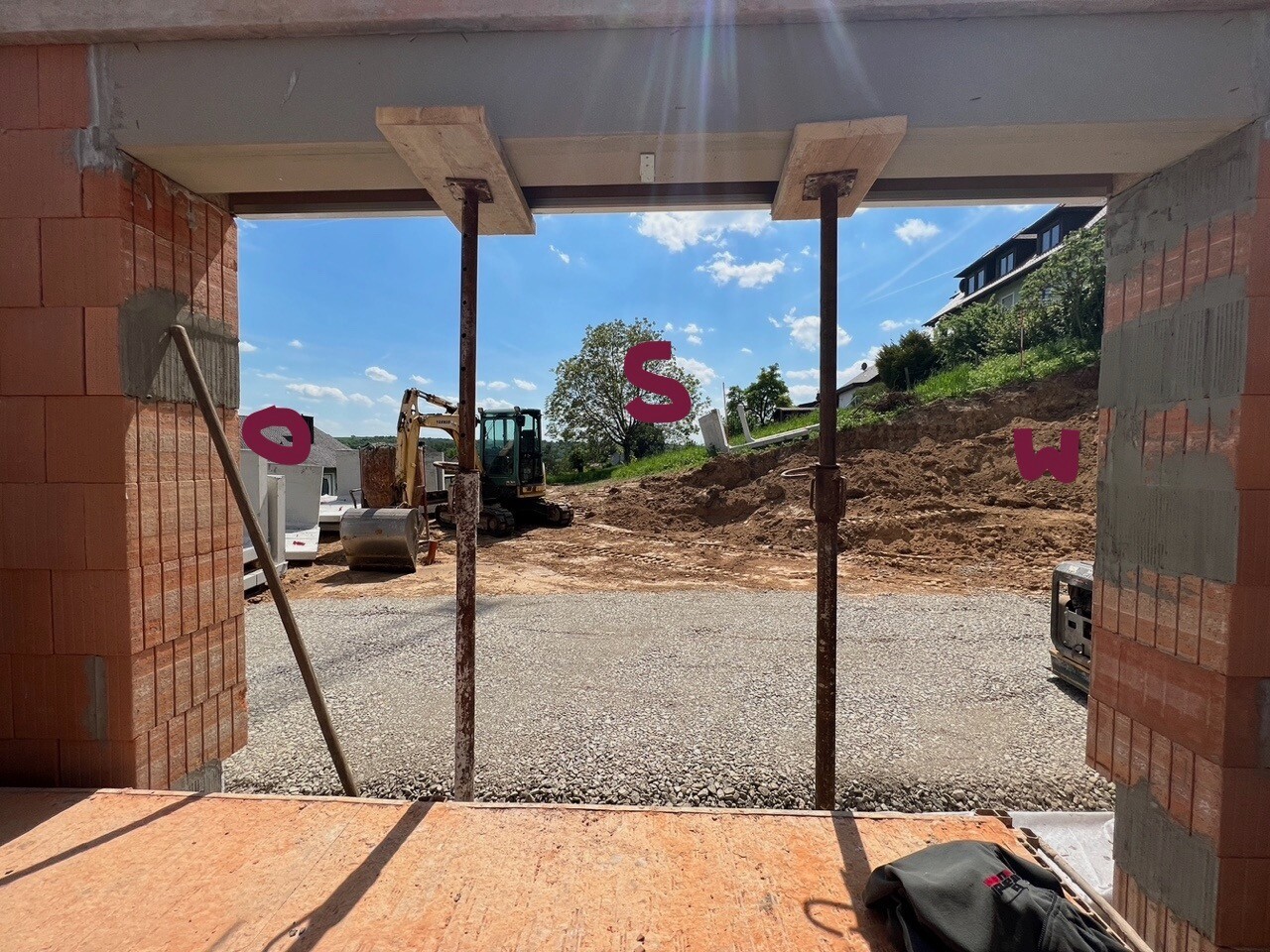 Offene Baustellenansicht mit roten Backsteinen, Stahlstützen, Bagger und Erdhügel vor Himmel.