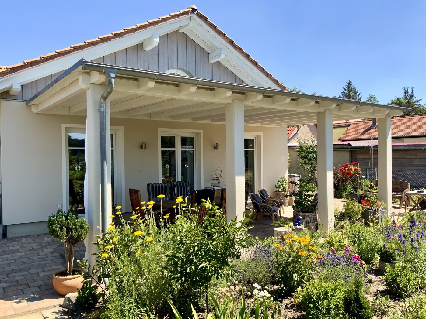 Heller Haus mit überdachter Terrasse und Sitzgelegenheiten; Garten mit bunten Blumen im Vordergrund.