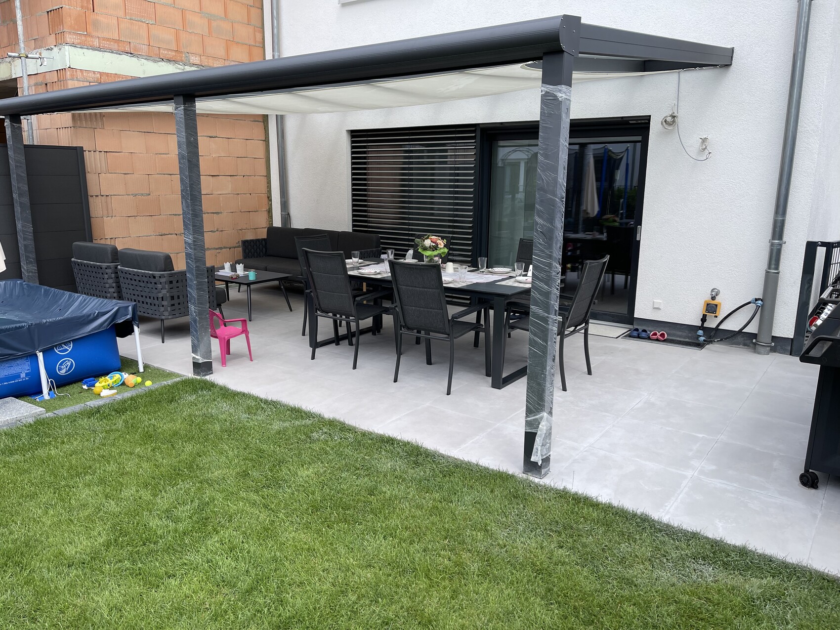 Gedeckter Gartentisch mit Stühlen unter einer Pergola auf der Terrasse; grüne Wiese daneben.