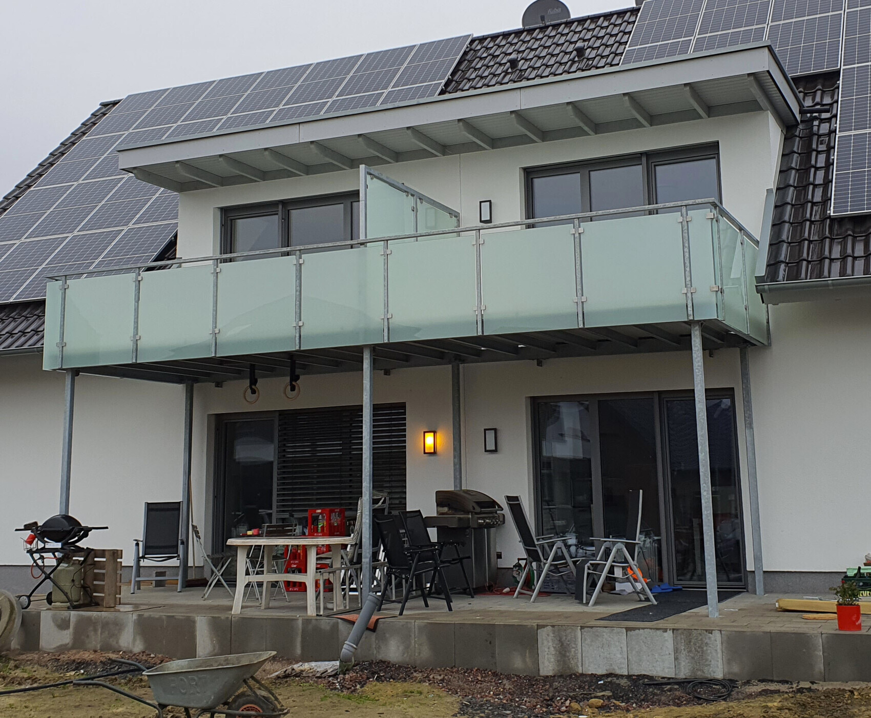 Modern house facade with balcony, glass railing, solar panels, and terrace with furniture.