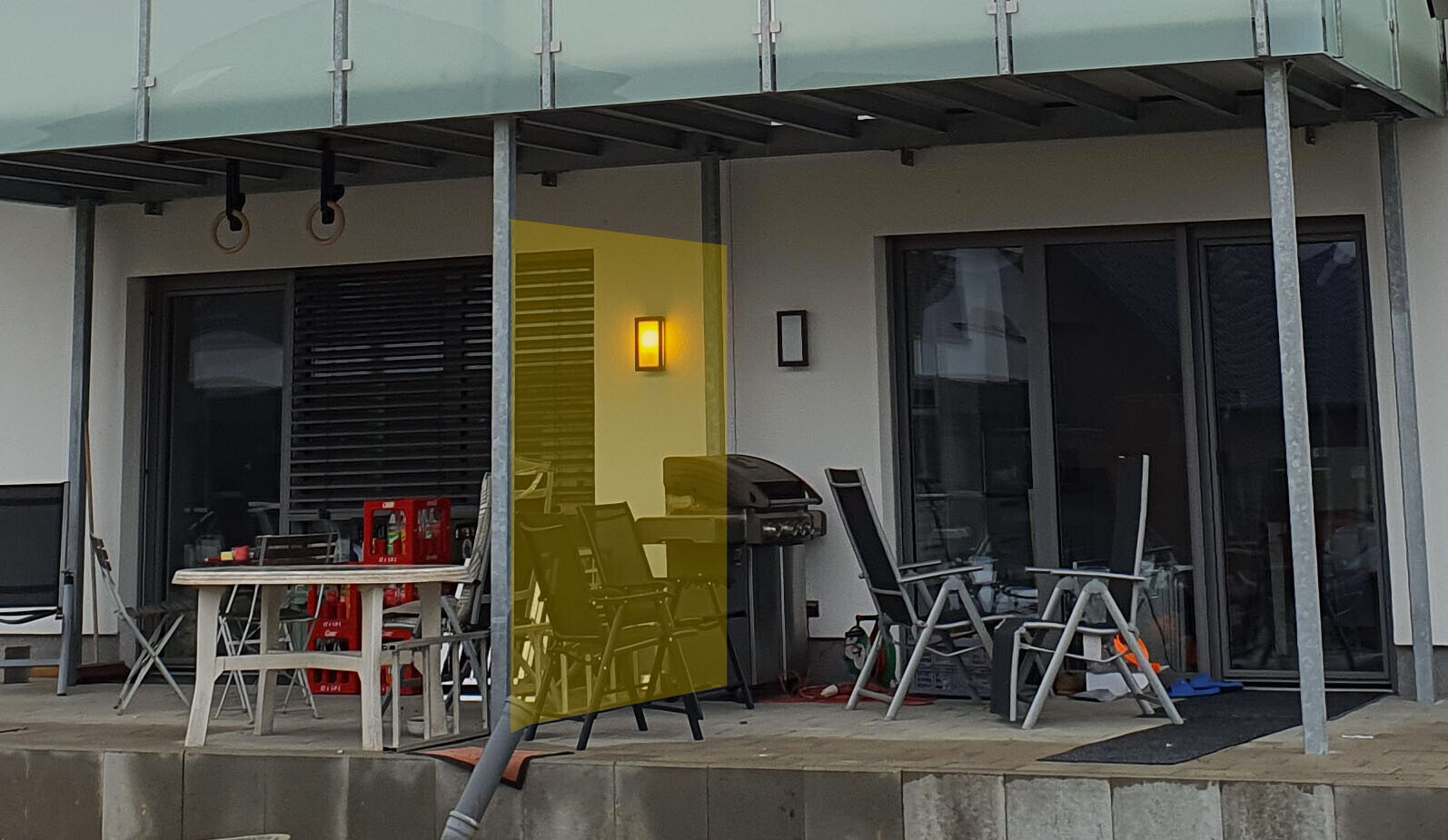 Terrace area in front of modern house: grill, outdoor table, chairs, glass doors, yellow tinted pane.