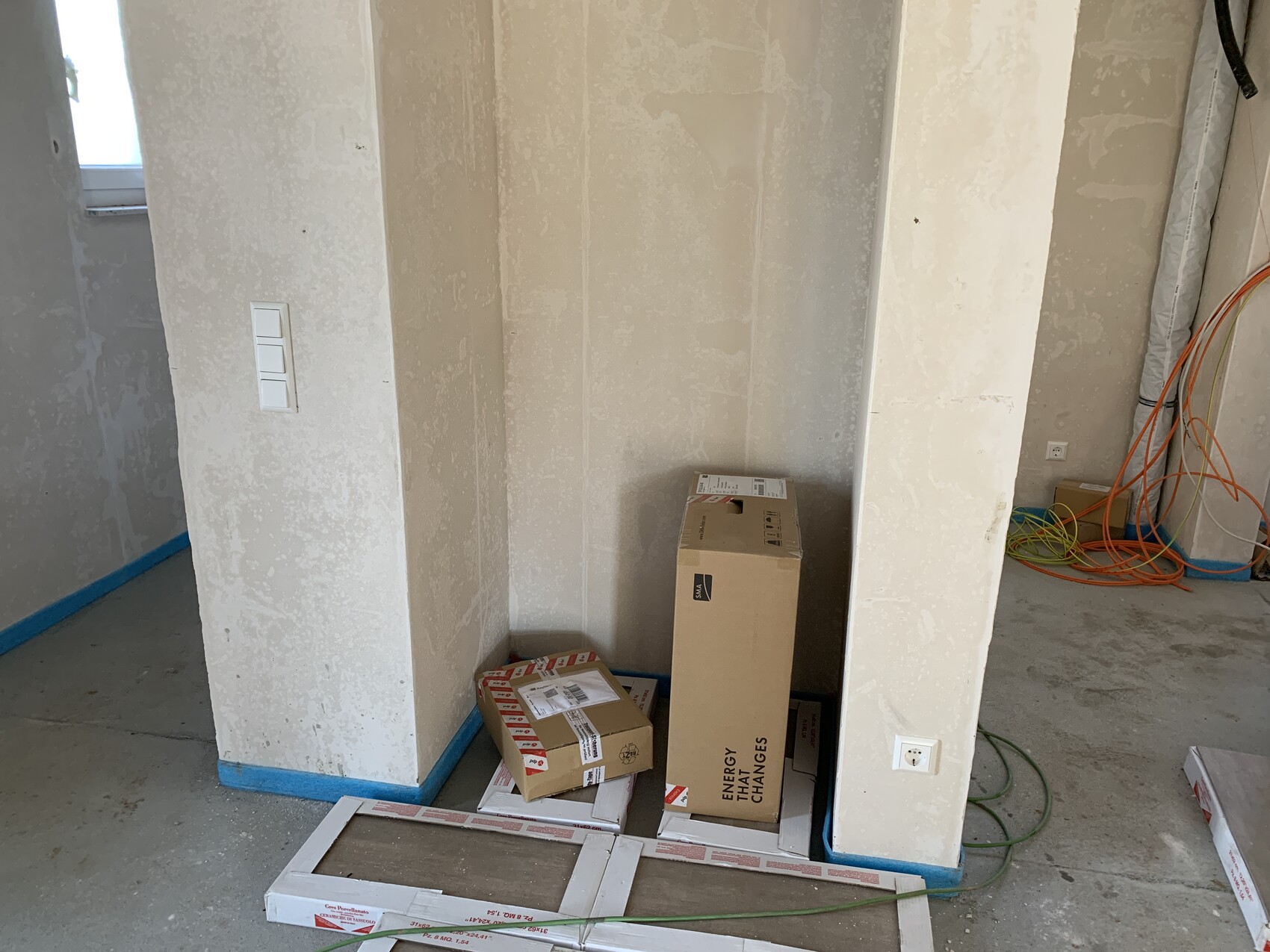 Baustelle: Unfertiges Zimmer mit verputzten Wänden, Kartons, bunten Kabeln und einer Steckdose.
