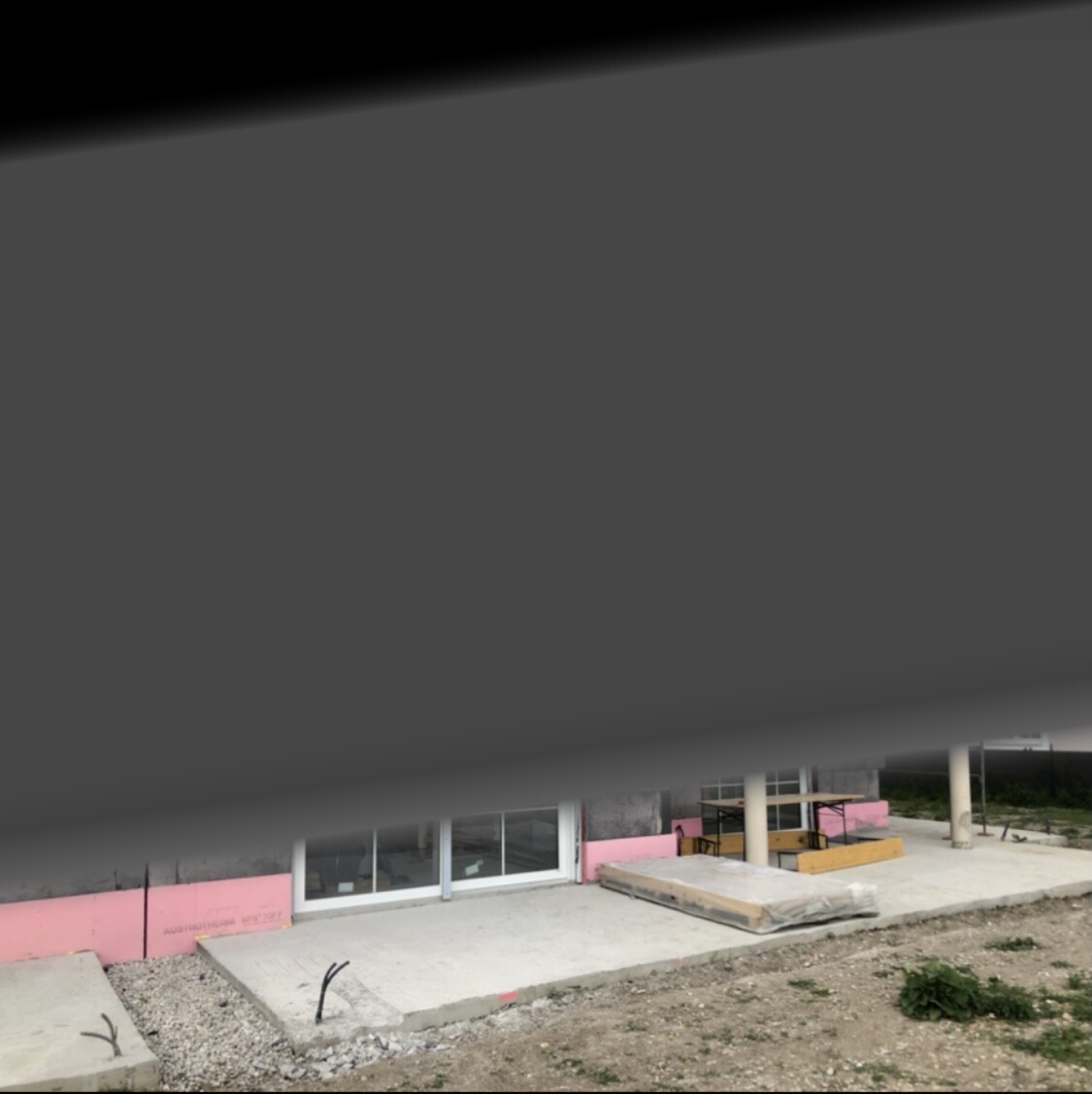 Construction site at the building: glass front, pink insulation panels, concrete floor and soil in front.