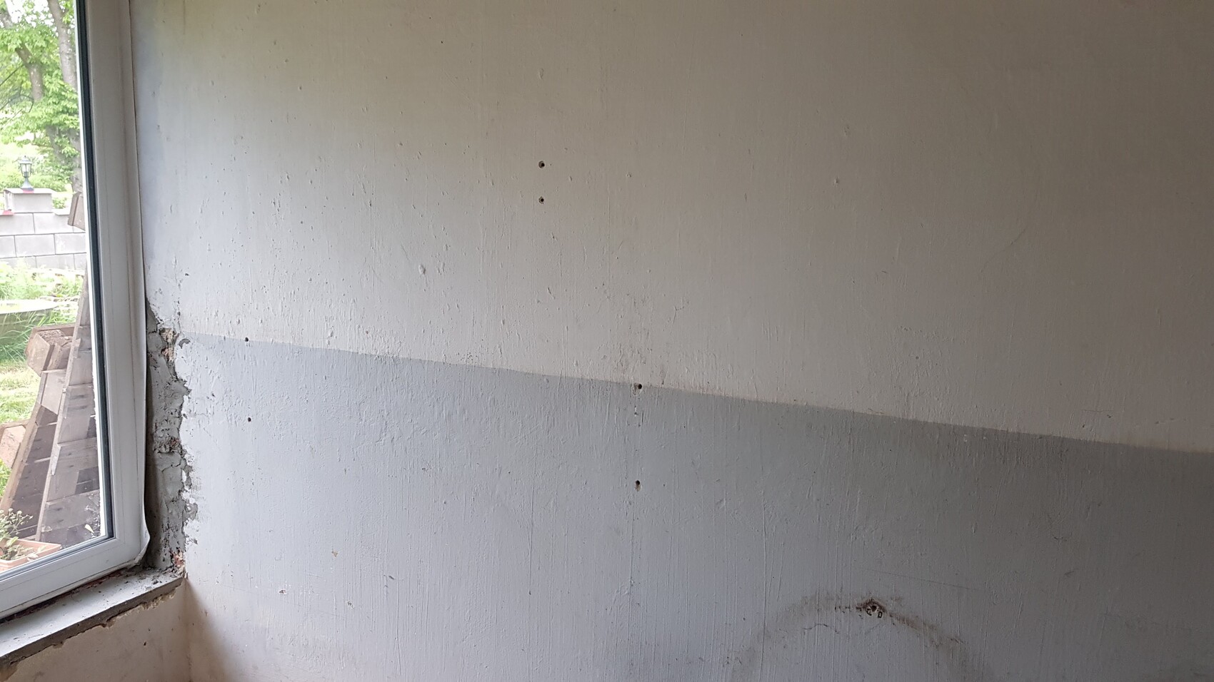 Light upper and gray lower parts of a wall with holes; window with outside view on the left.