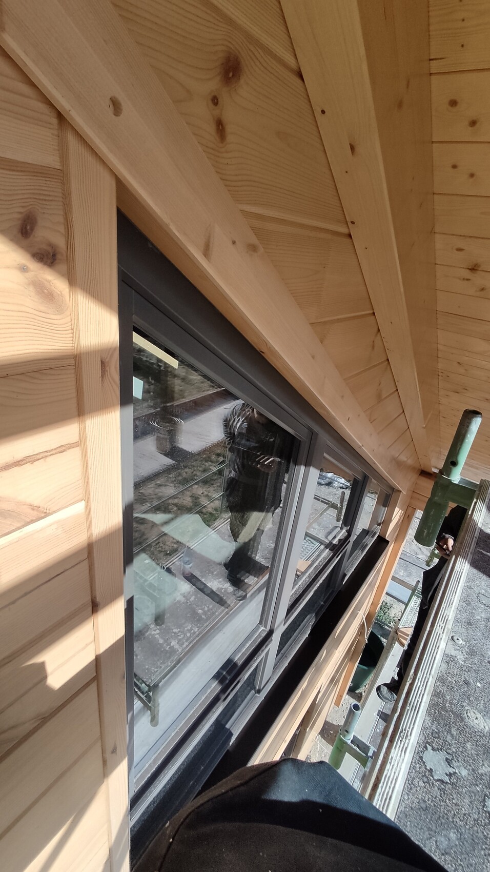 Außenansicht eines Holzhauses im Bau; Fensterrahmen, Holzfassade und Gerüst sichtbar.