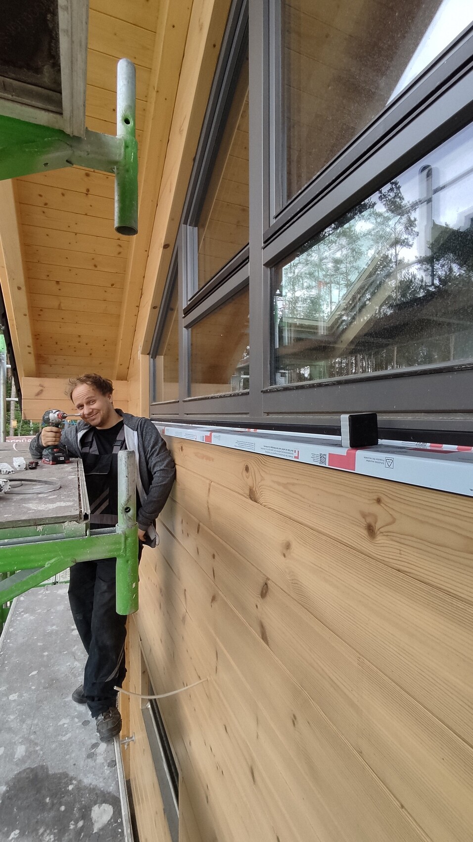 Mann mit Akku-Bohrschrauber bohrt Holzfassade neben Fenstern; Baugerüst sichtbar.