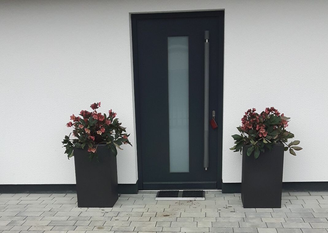 Moderne Haustür in Dunkelgrau, flankiert von zwei hohen Pflanzkübeln mit rosa Blüten.