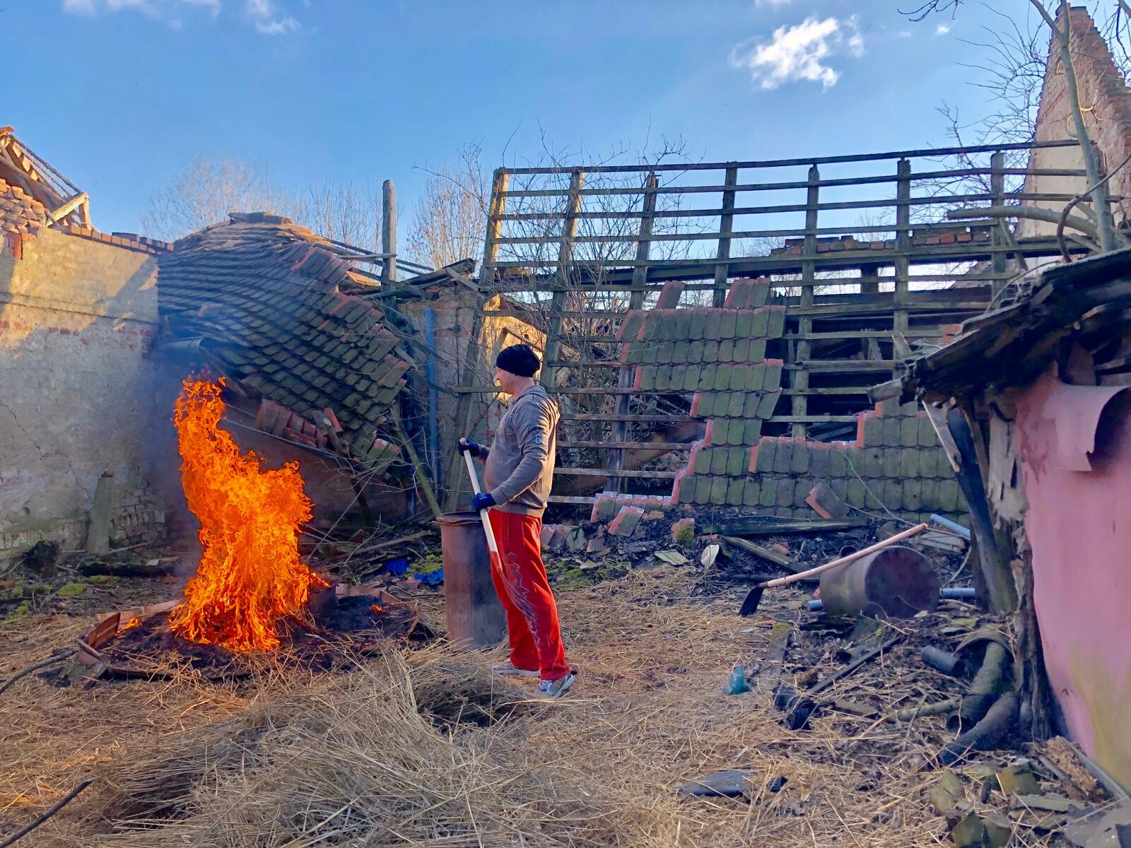 Man in gray hoodie and red trousers standing next to a burning fire in ruined brick ruins.