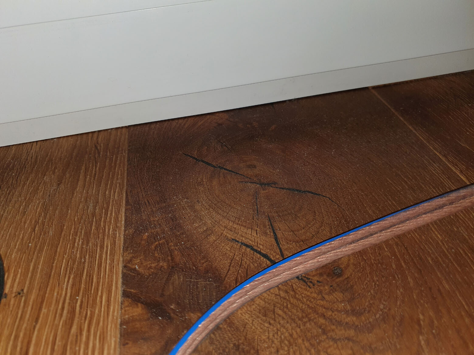 Wood floor with dark scratches and cracks, white baseboard; blue cable lying on top.
