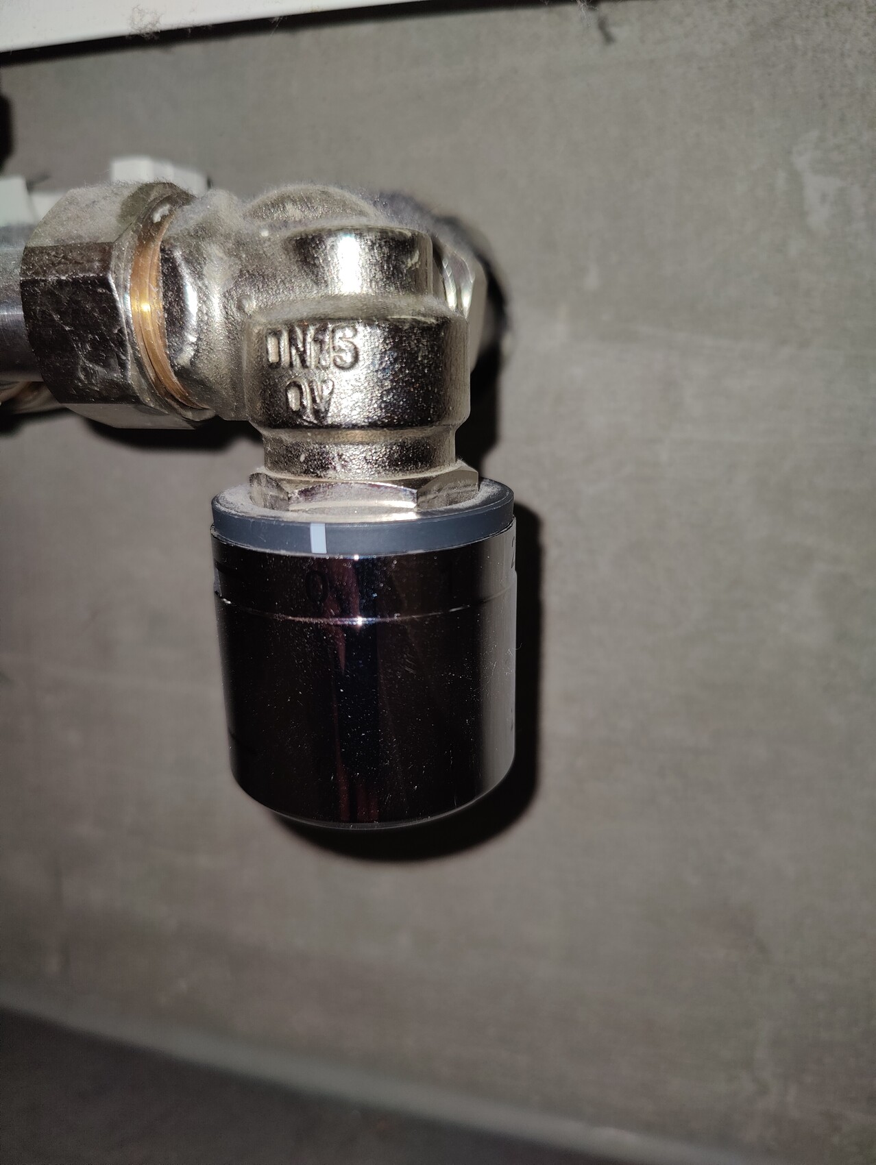 Close-up of a metal pipe connector with valve and black cylinder, concrete wall in the background.