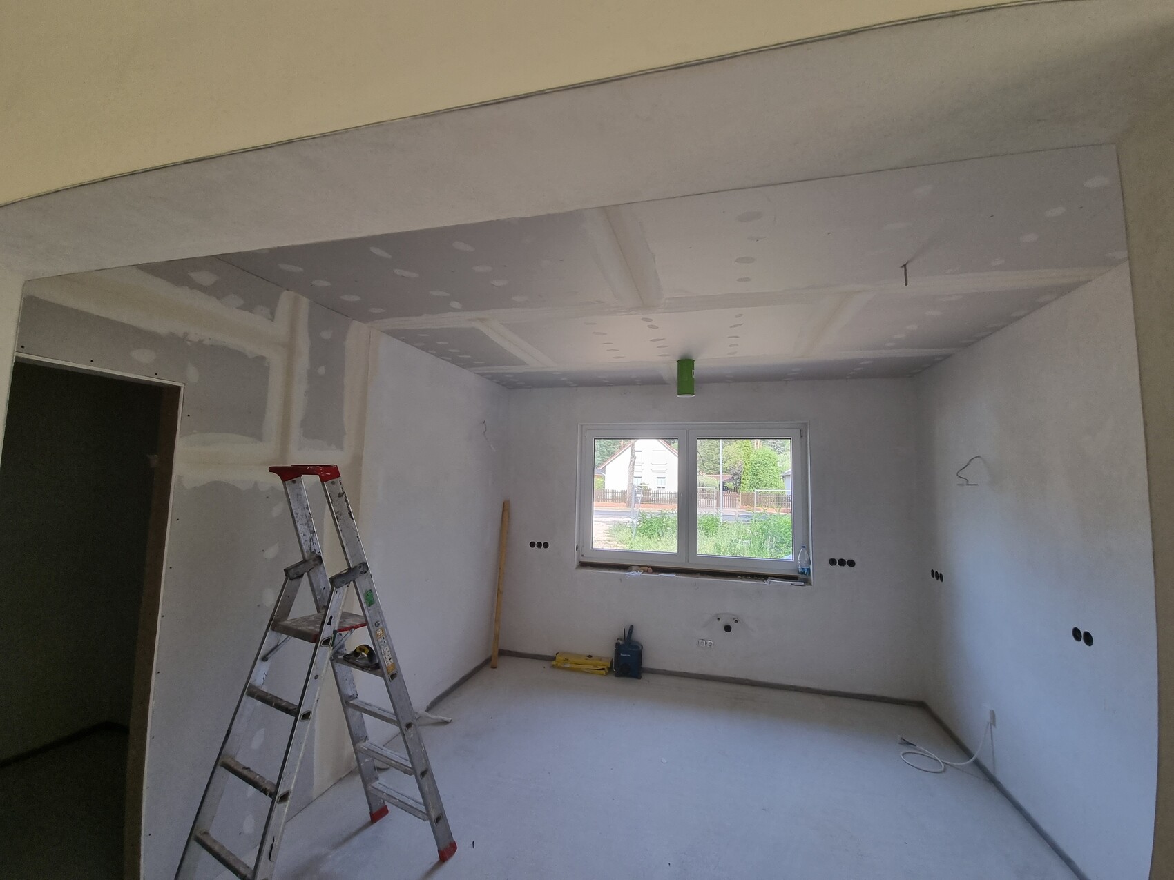 Rohbauzimmer: gespachtelte Wände/Decken, eine Leiter, Werkzeuge und ein Fenster mit Außenblick.