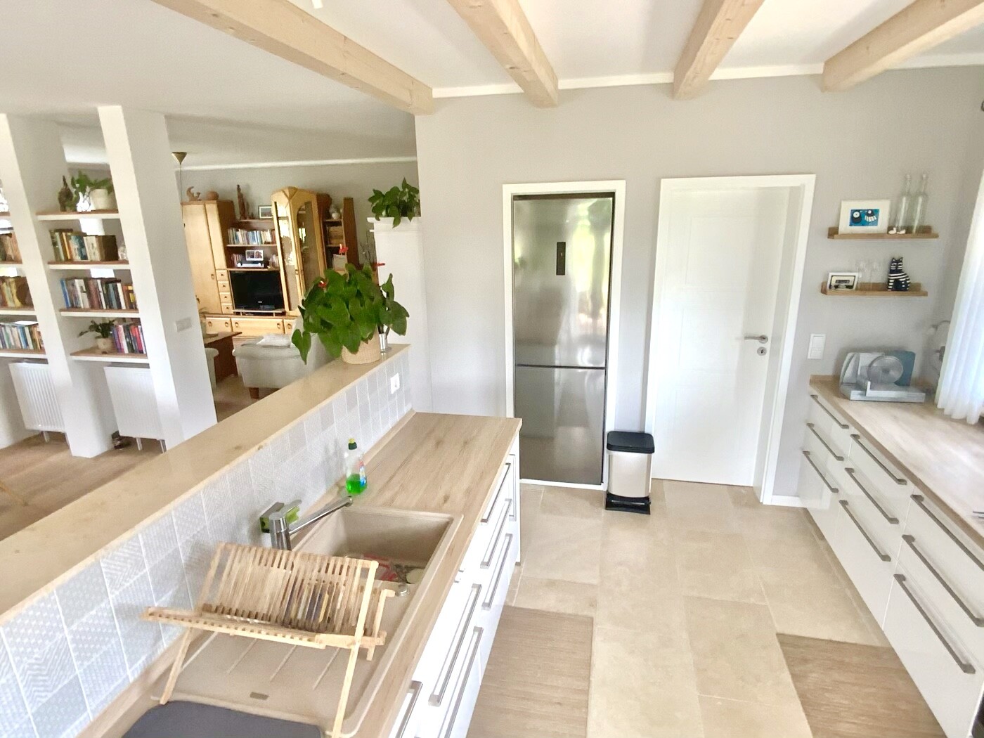 Open kitchen with wooden beam ceilings, sink on the island, plant, and bookshelf in the background.