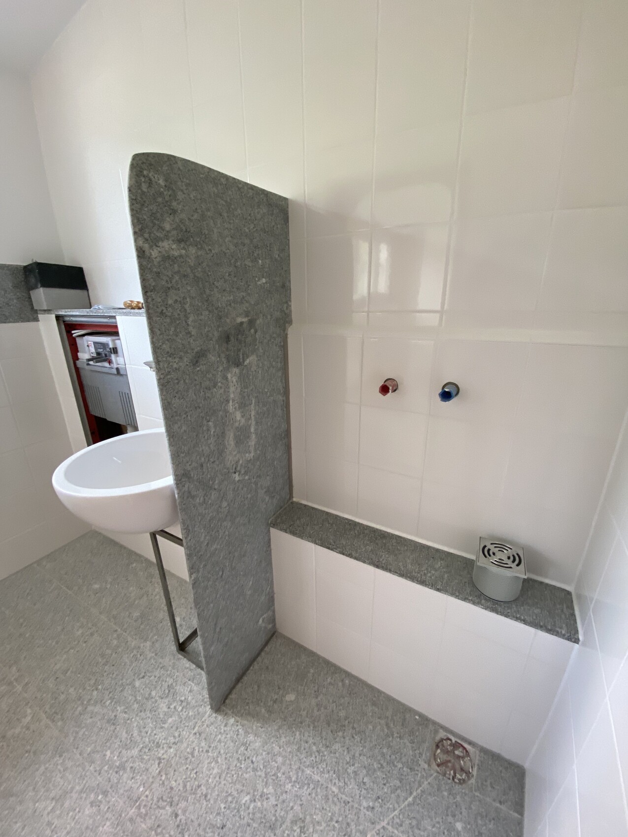 Sink in front of grey granite partition wall, red and blue water taps, grey floor.