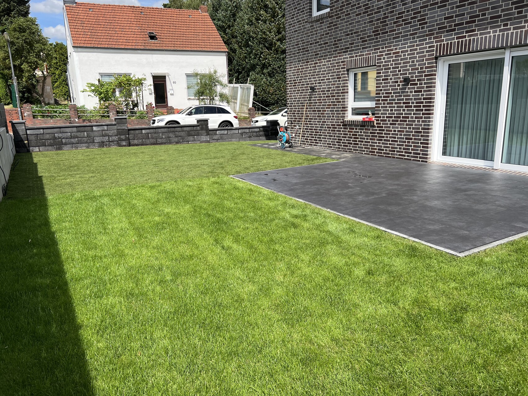Garden with green lawn, dark terrace floor on the brown brick house; parked car visible.