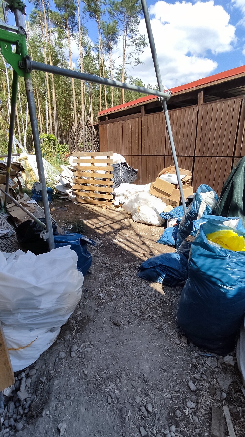 Outdoor area with garbage bags, pallets, cardboard and construction waste next to wooden wall; forest in the background.