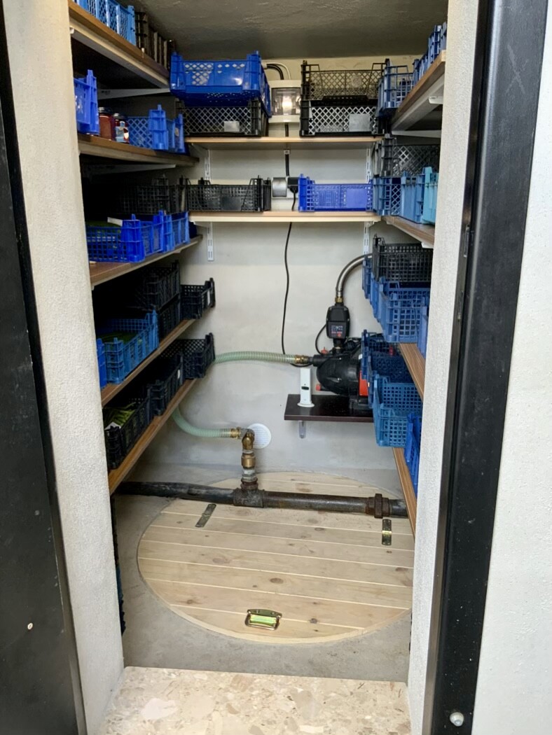Small storage room with shelves full of blue plastic containers; pump, pipes, and hose on the floor.