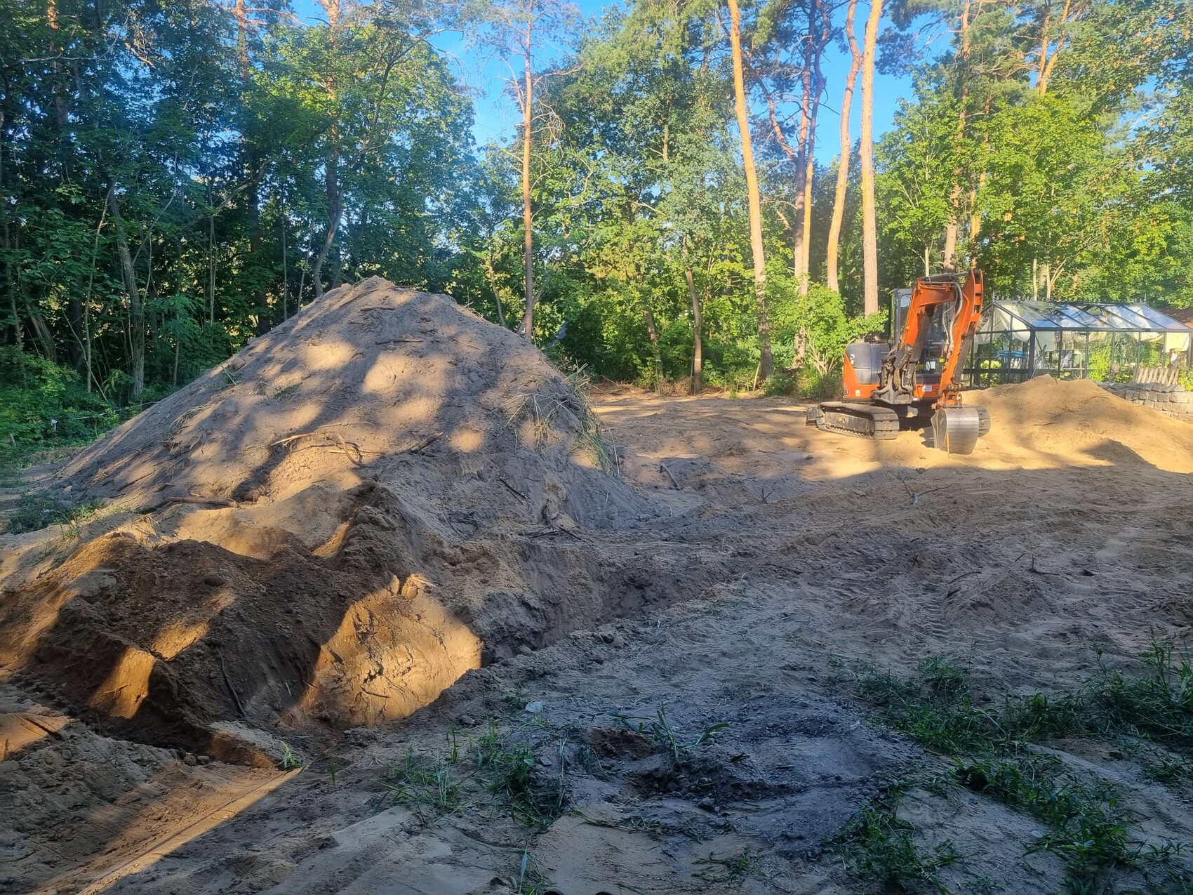 Großer Sandhaufen auf einer Baustelle, orange Bagger rechts, Wald im Hintergrund
