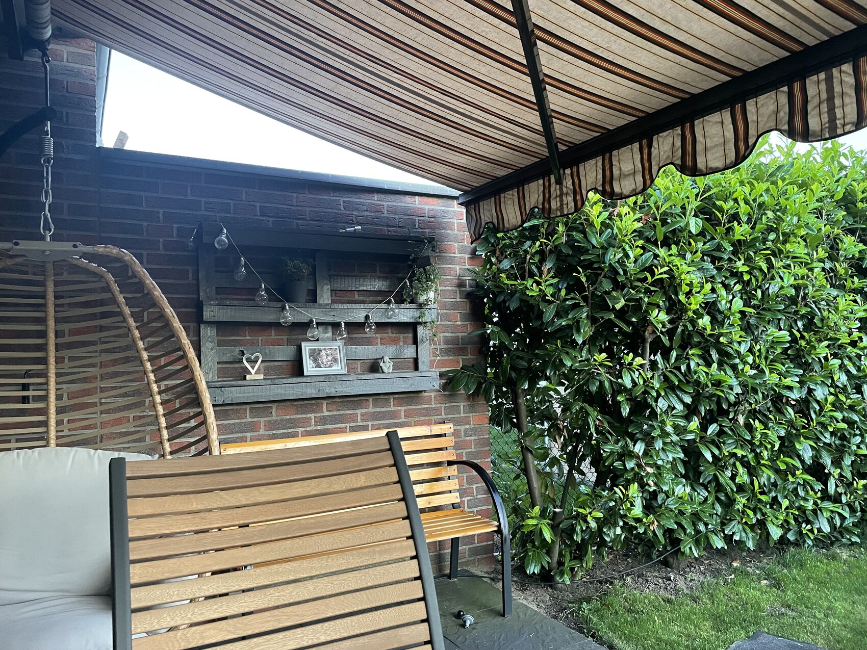 Cozy terrace with wooden benches, hanging chair, string lights, decorative shelf on brick wall, hedge.