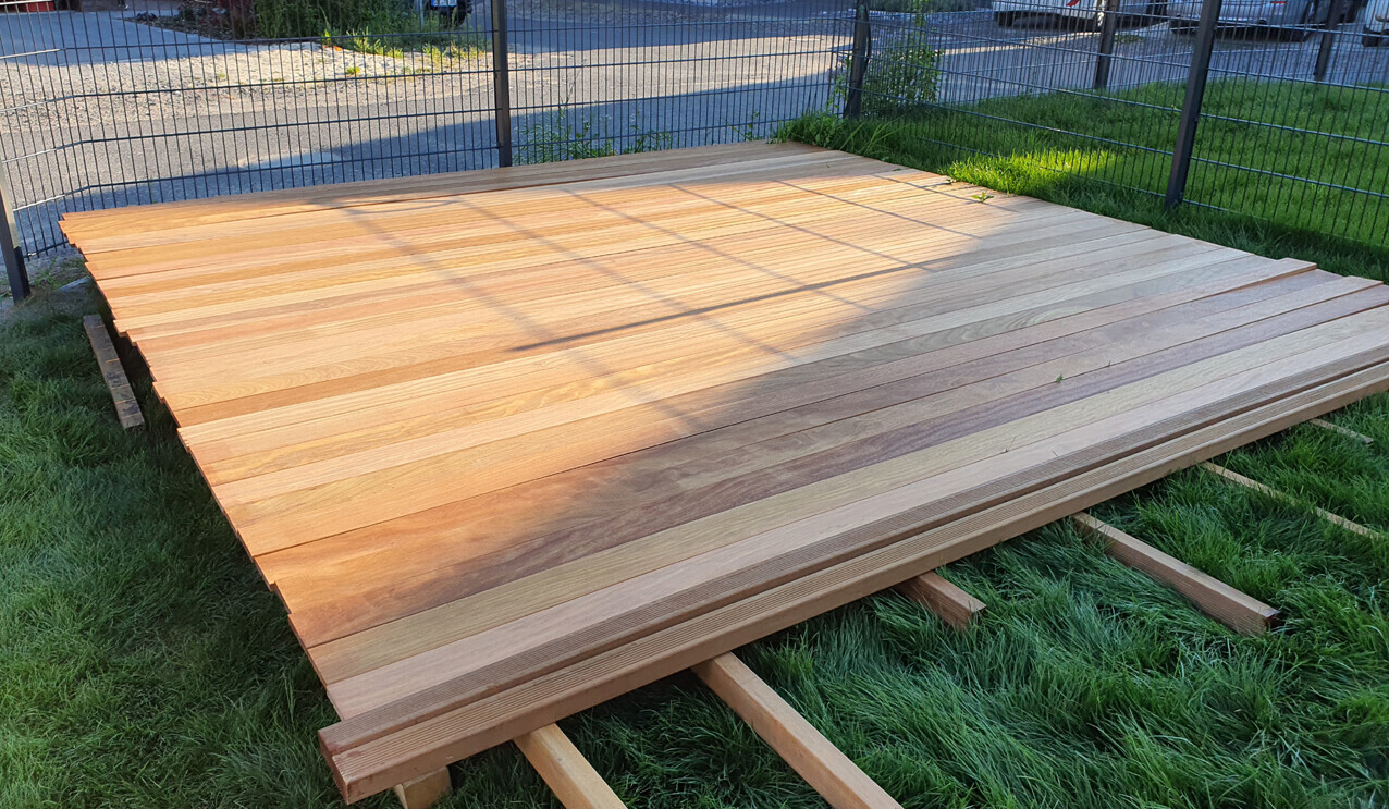 Große Holzplattform aus quer verlaufenden Dielen liegt auf Holzbalken auf Gras neben einem Zaun.