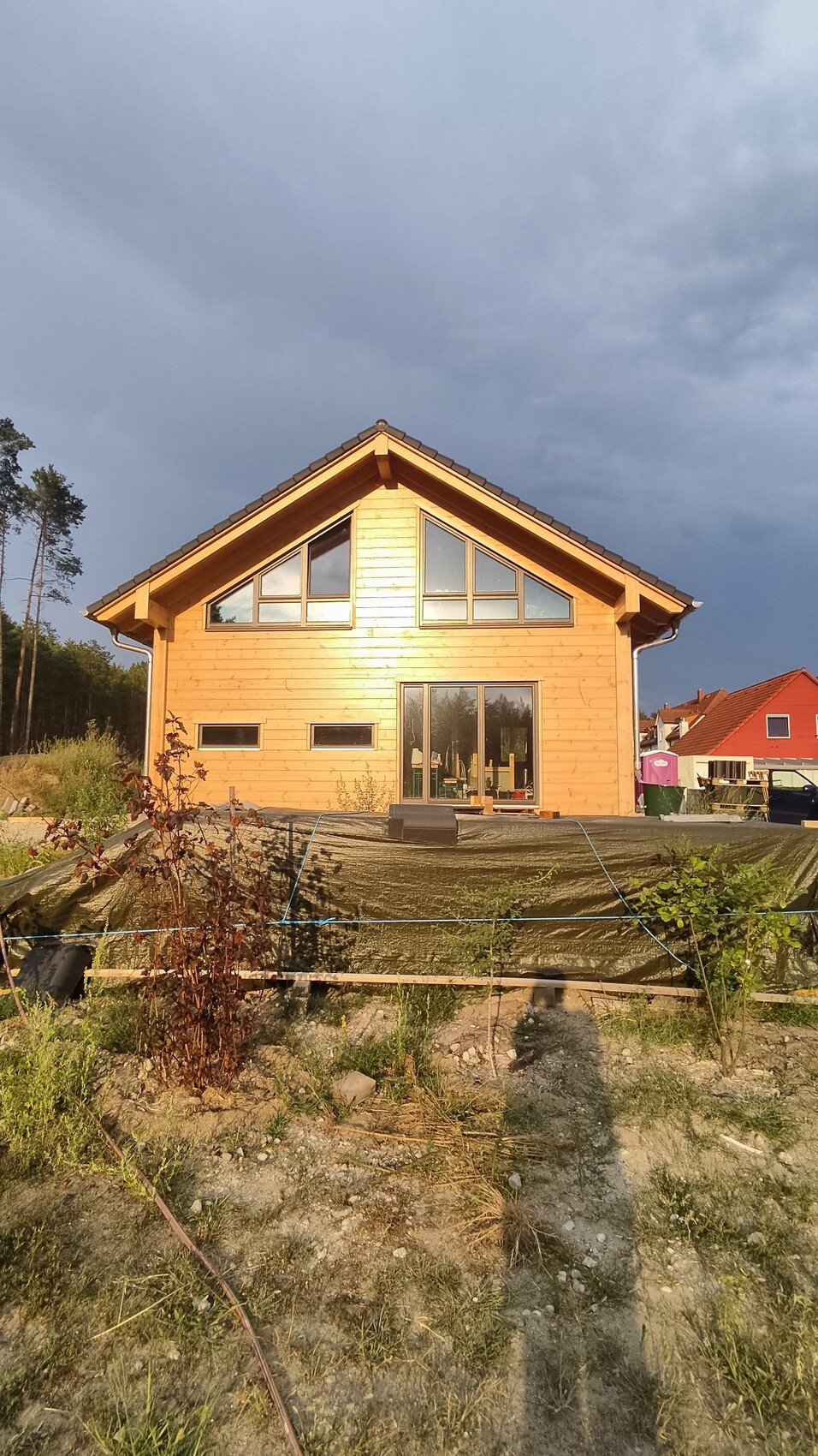Wooden house with large triangular gable windows, construction area with tarpaulin in front, dark sky