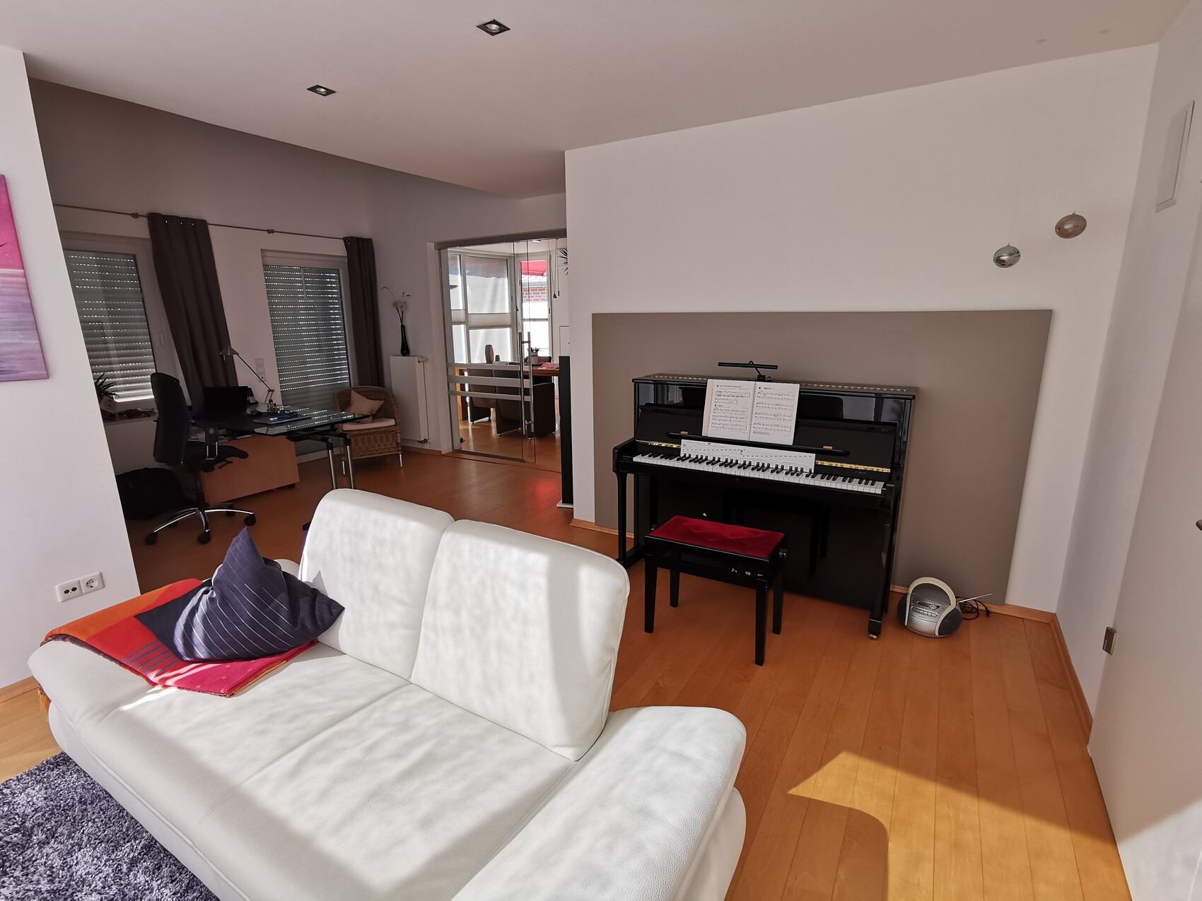 Living room with white sofa, black piano, sheet music, workspace on the left, wooden floor.