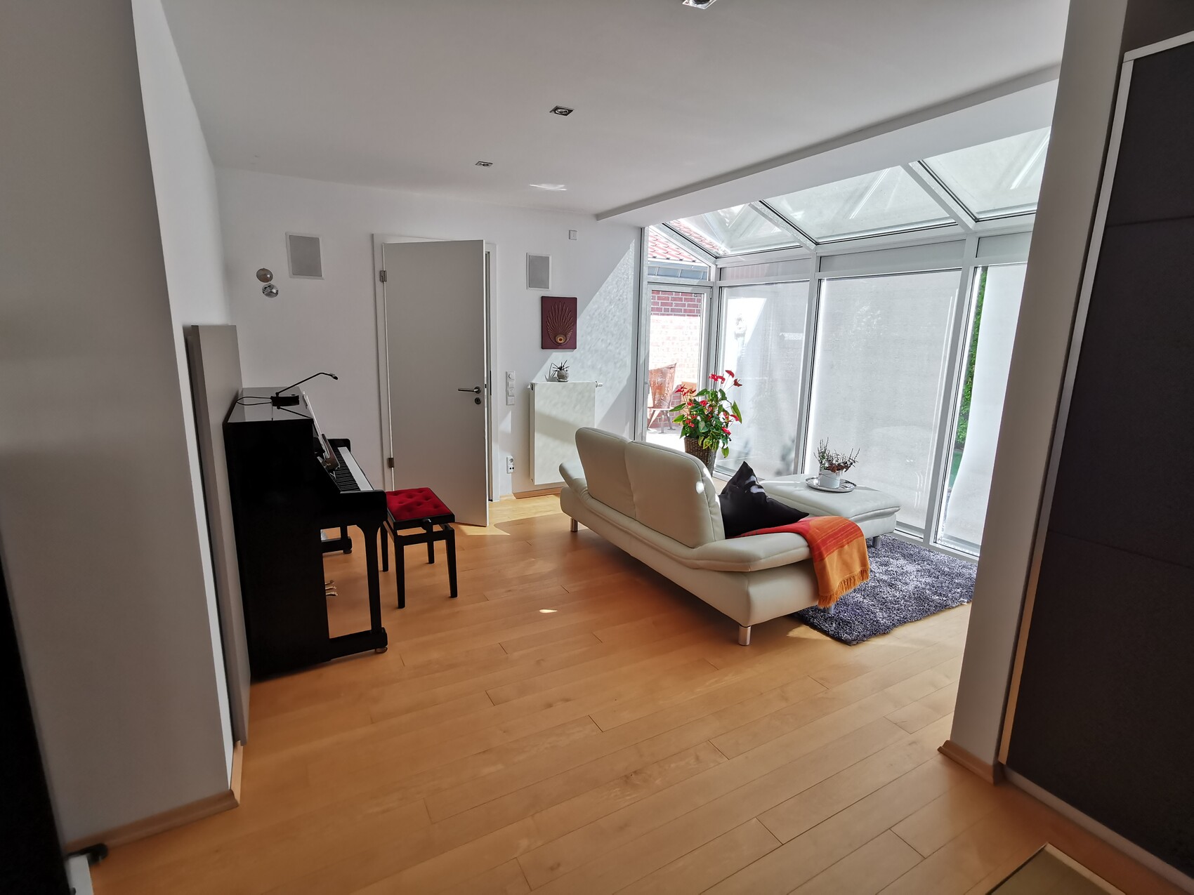 Bright living room area with white leather sofa, wooden floor, piano on the left, and glass sunroom.