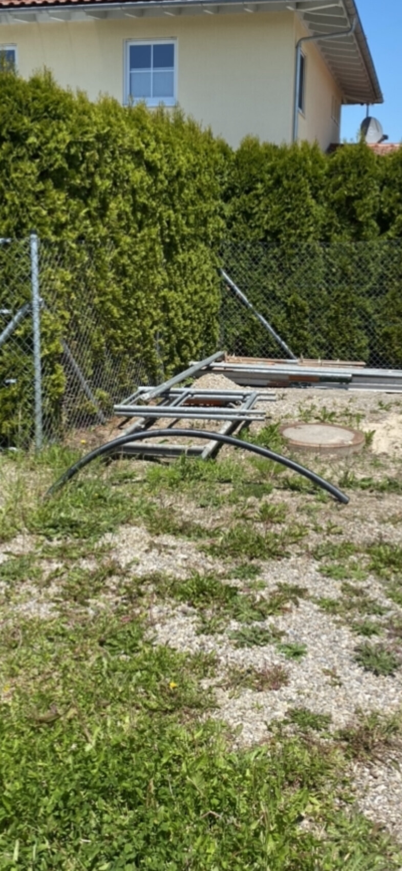 Baustellenbereich mit Metallrohren und Gestell vor grünem Zaun neben dem Haus.