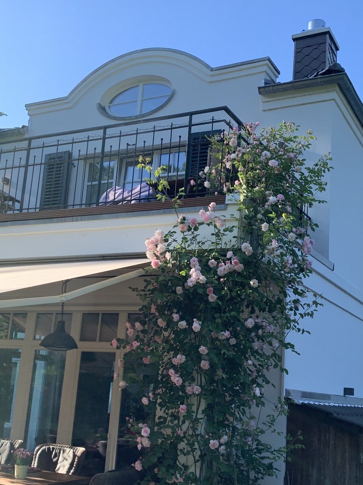 White house facade with balcony and rounded upper window, pink roses on the wall; terrace with furniture.