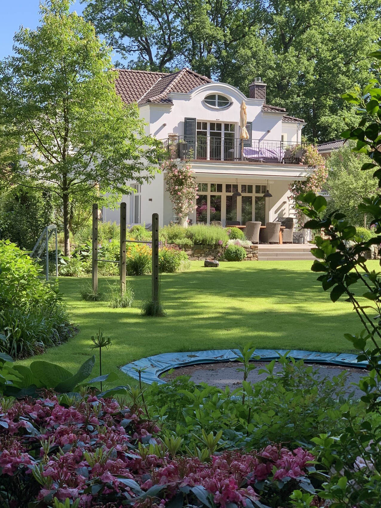White two-story house with balcony, terrace and garden; trampoline in the foreground and flower beds.