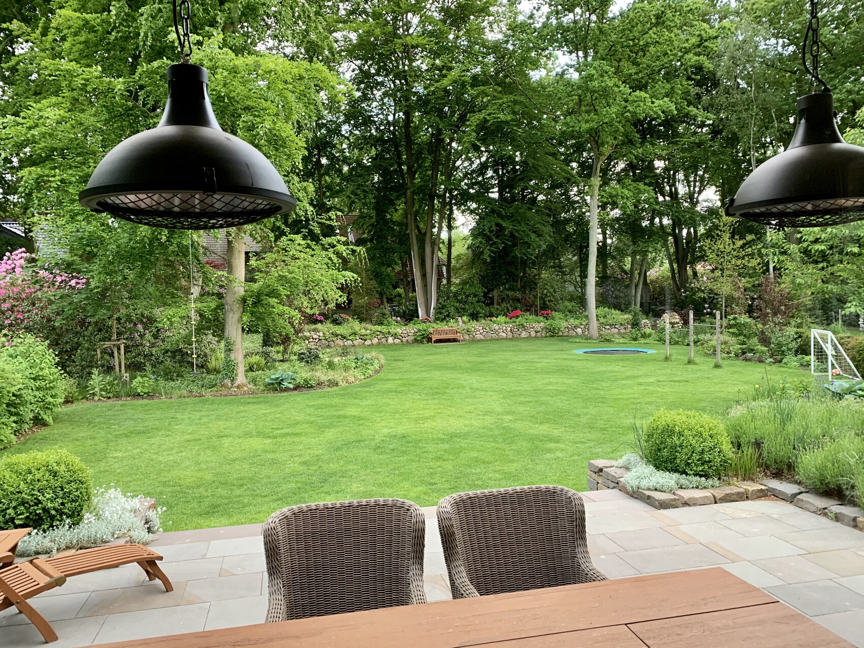 Garden view: terrace with table and wicker chairs in the foreground, green lawn, trees and beds.