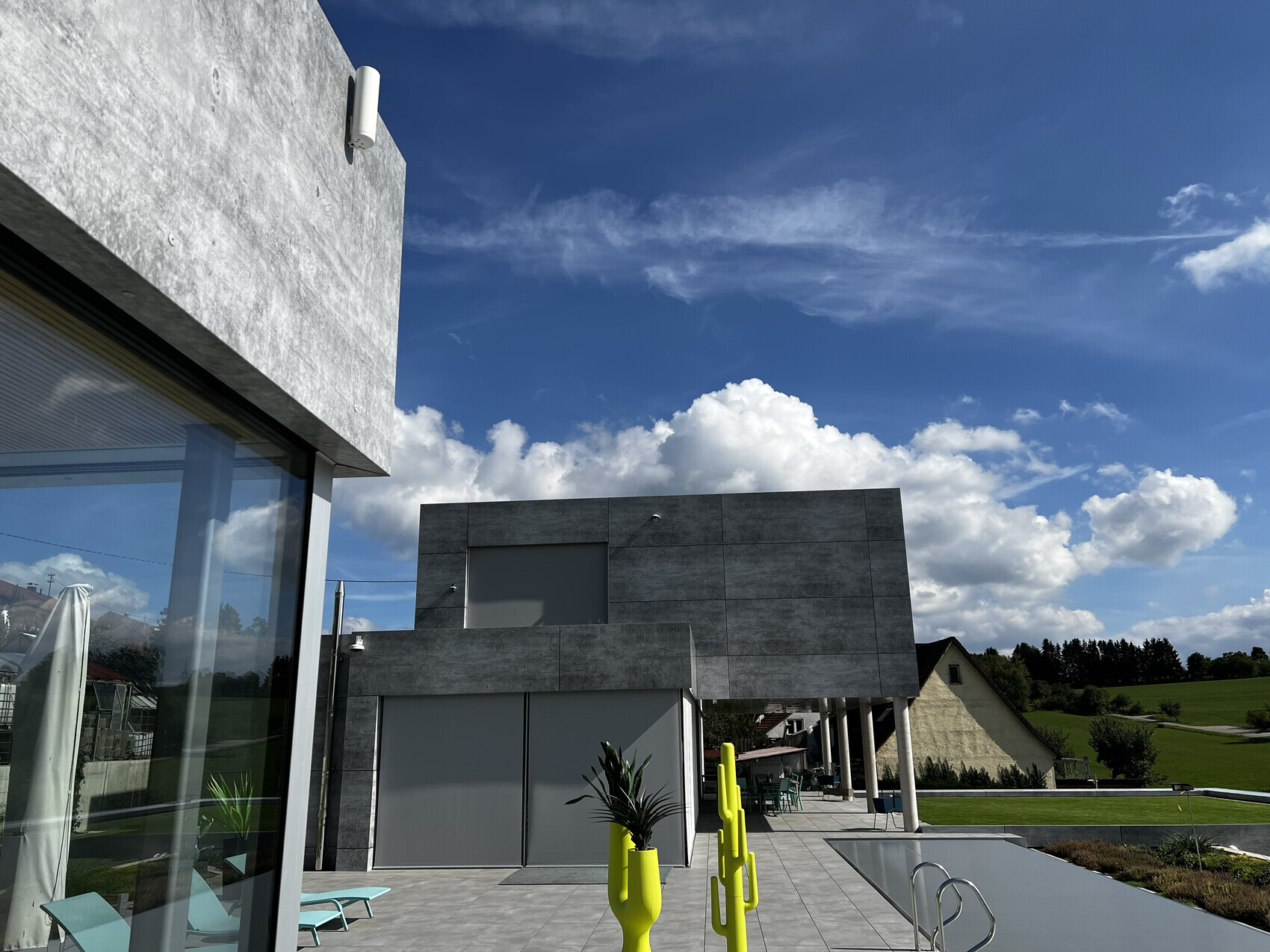 Moderne graue Architektur mit Glasfront Terrassenbereich Pool und gelbe Stäbe vor grünem Gelände