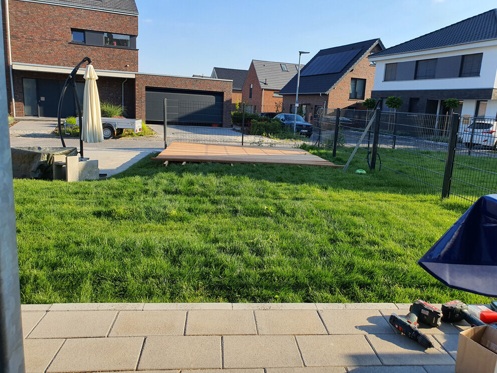 Garten mit grünem Rasen, Holzdeck und Werkzeug auf Terrasse, Häuser im Hintergrund.