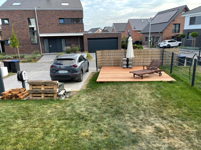 Suburban garden with wooden deck, lounge chairs, sun umbrella, parked car, pallets on the left.