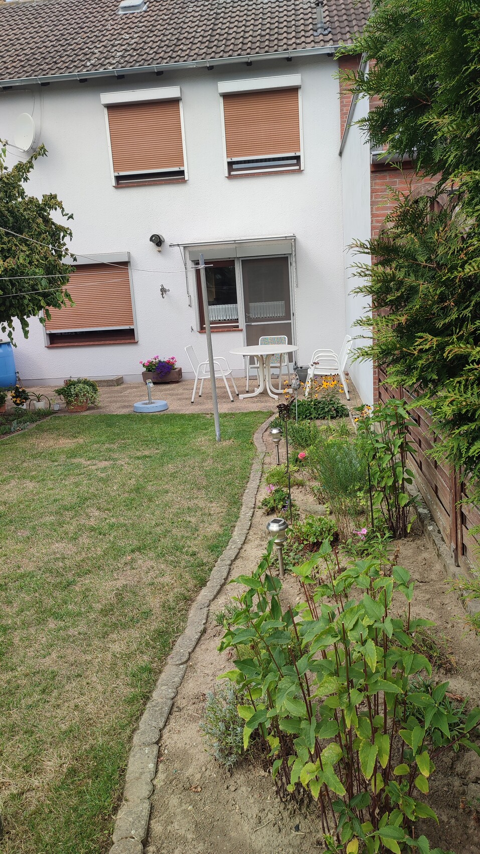 Garden behind a white house: patio with table and chairs, flowerbeds, lawn, fence.