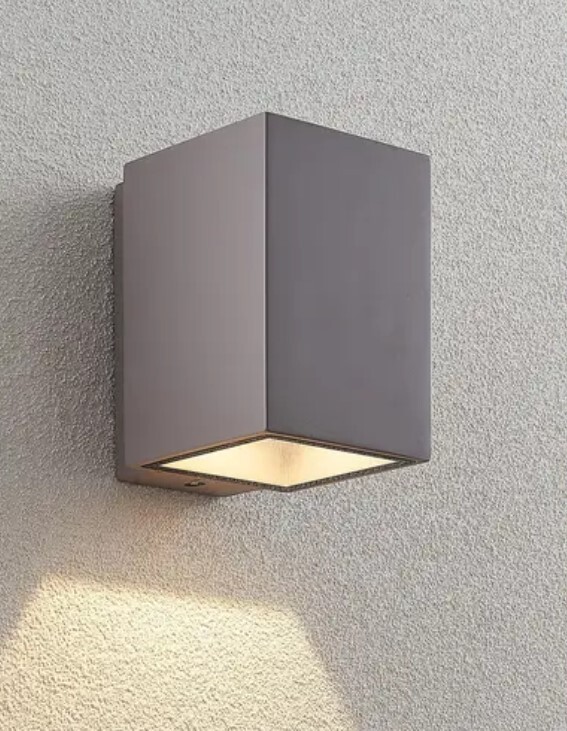 Outdoor wall light in a gray square cube shape, shining downwards on a rough wall.
